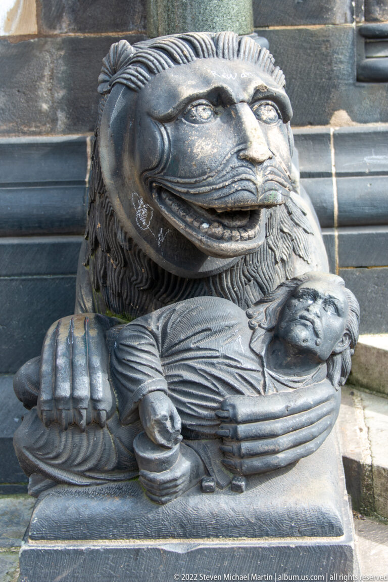 Close-up of a dark stone lion carving with a smaller figure held beneath its front paws.