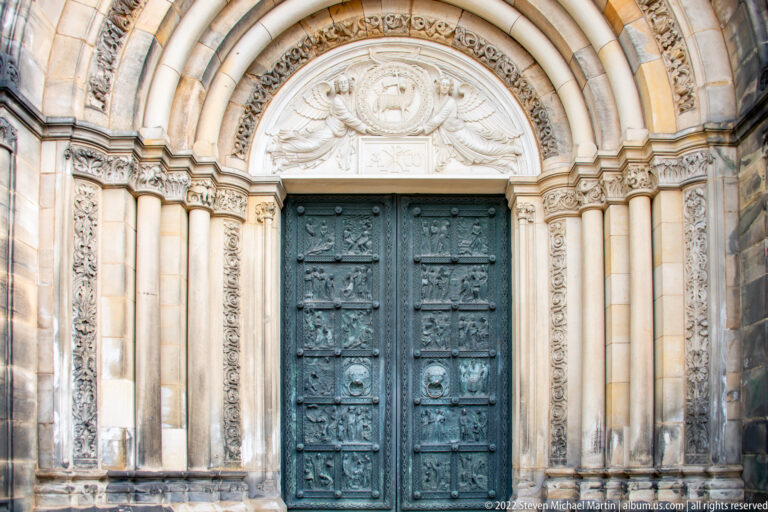Ornate round-arched cathedral portal with carved stone surrounds and dark bronze doors covered in relief panels.