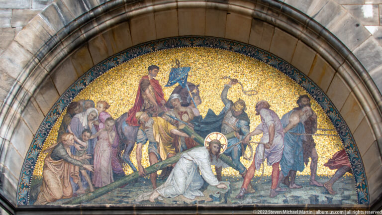 Gold-ground arched mosaic of Christ carrying the cross, surrounded by robed figures at a cathedral entrance.