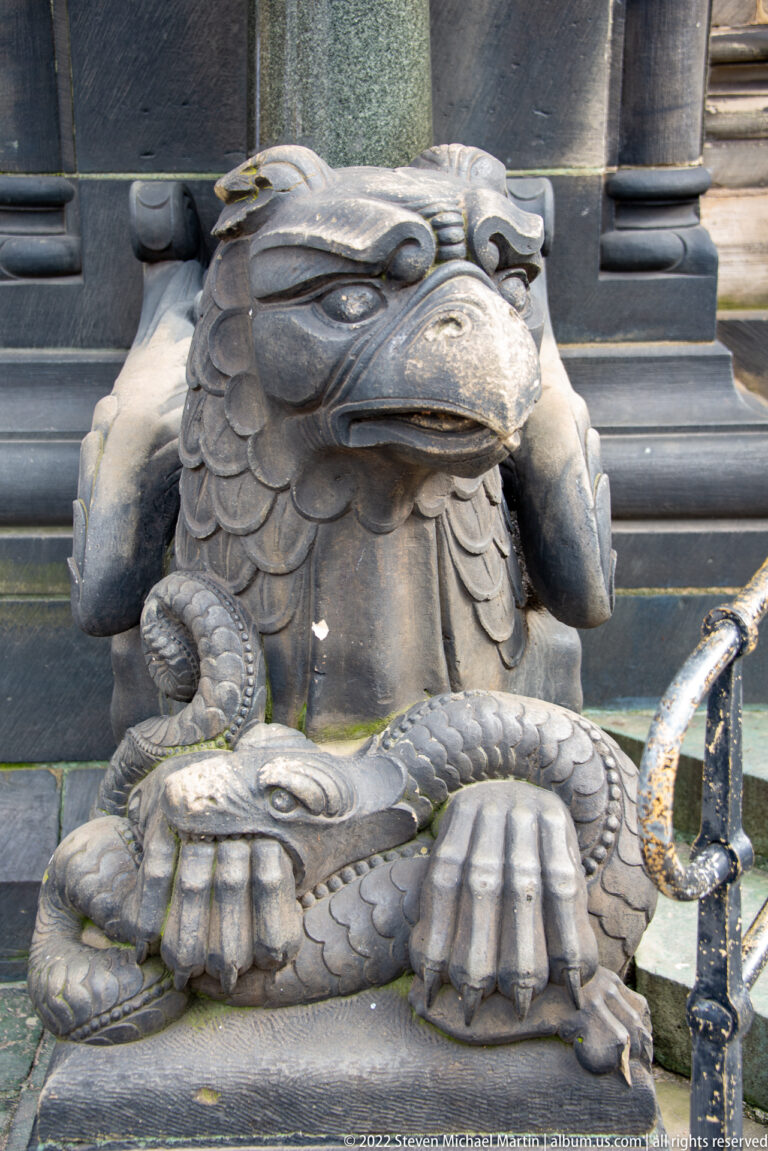 Dark stone griffin-like creature with hooked beak, feathered head, large paws, and a smaller coiling animal beneath.