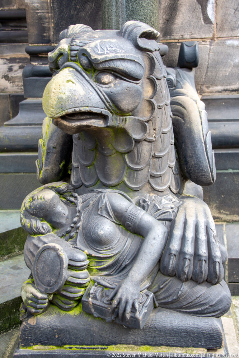 Weathered griffin-like stone sculpture with hooked beak, feathered head, heavy paws, and green surface growth.