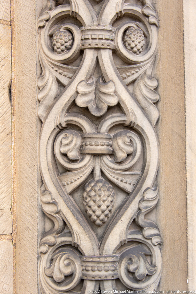 Vertical pale-stone relief panel with symmetrical scrolls, leaf motifs, circles, and a central diamond form.