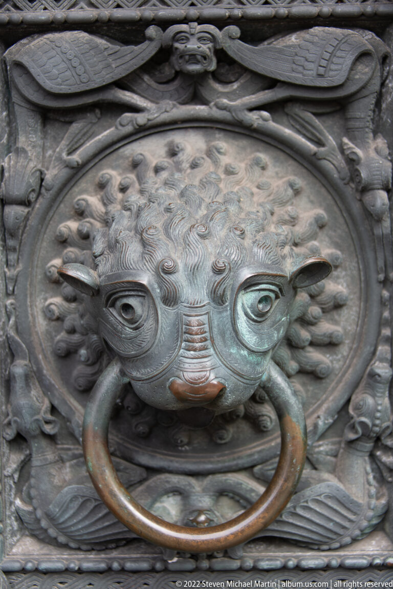 Bronze lion-head door knocker holding a large circular ring, set within an ornate metal plate.
