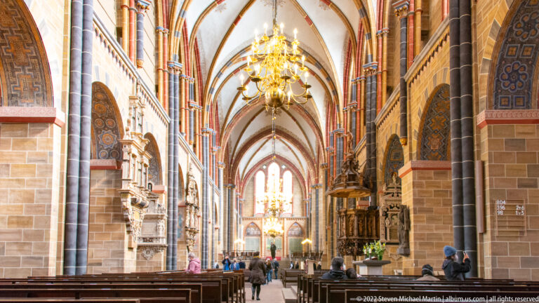 Long central aisle of Bremen Cathedral beneath pointed vaults, with chandelier, pews, monuments, and distant sanctuary.