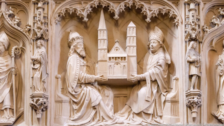 Two mitred clerics in carved stone hold a model twin-towered church beneath ornate Gothic canopies.