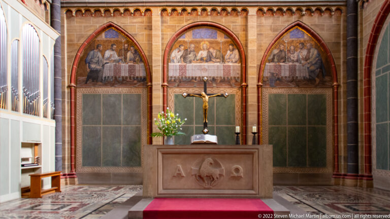 Stone altar with central crucifix before three arched painted panels depicting meal scenes inside Bremen Cathedral.