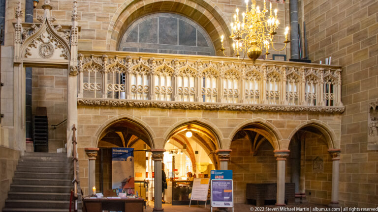 Carved Gothic stone gallery above pointed arches and a staircase inside Bremen Cathedral, with visitor facilities below.