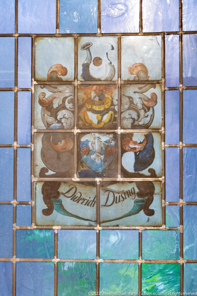Heraldic stained-glass panel with shield, helmet, scrolling mantling, and the painted name David Dusing.