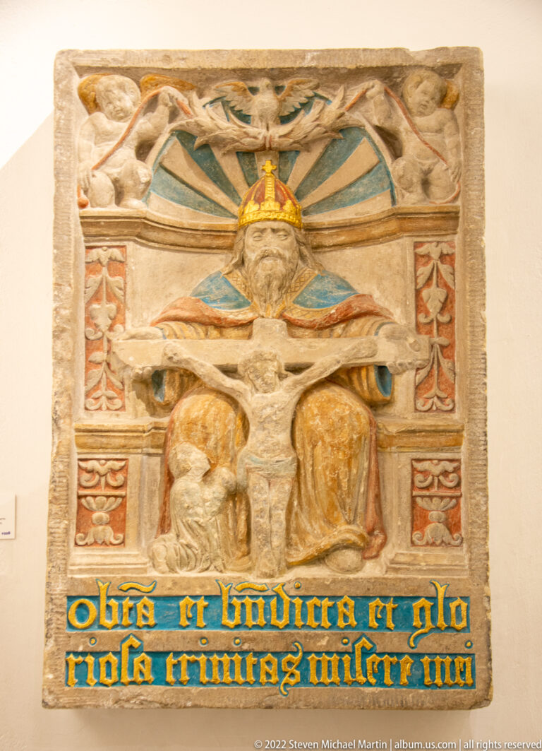 Painted stone relief with crowned bearded figure above two robed figures and a blue Latin inscription band.
