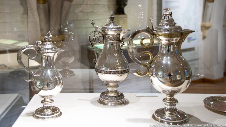 Three ornate historic silver ewers with handles and lids displayed together in a museum case.