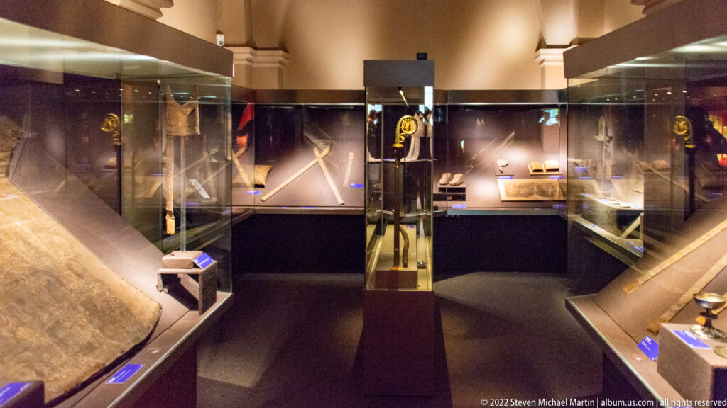 Dark museum gallery with glass cases displaying small historic artifacts and metalwork beneath vaulted architecture.