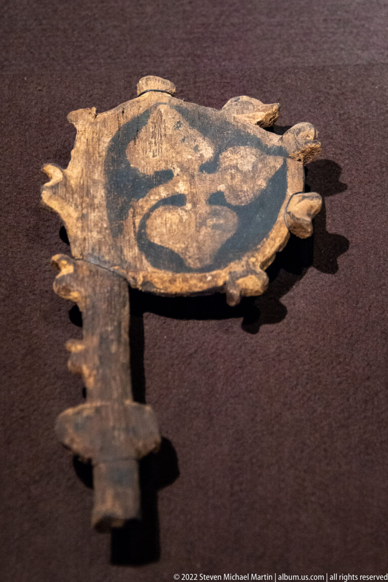 Large old iron key with broad decorated bow containing a painted heraldic motif, displayed in a museum case.
