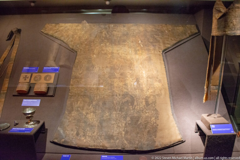Large faded historic textile with indistinct figures and decorative border hanging vertically in a museum display.