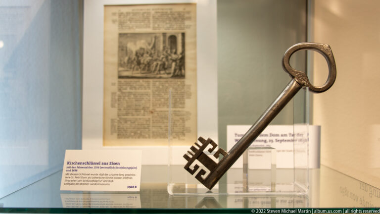 Large historic iron key with circular bow and heavily notched bit displayed upright in a museum case.