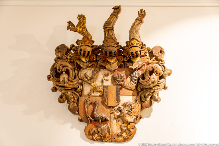 Ornate polychrome heraldic plaque with multiple shields, helmets, crests, and scrolling decoration.