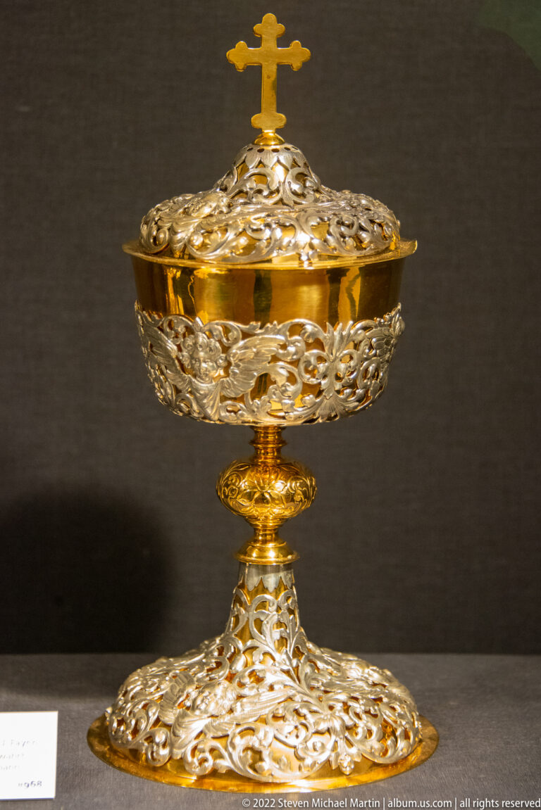 Ornate gilded openwork liturgical vessel on a stem and broad foot, topped by a small cross.