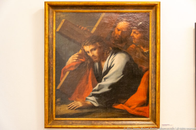 Close religious painting of Christ bowed under the cross with two figures pressing behind him.