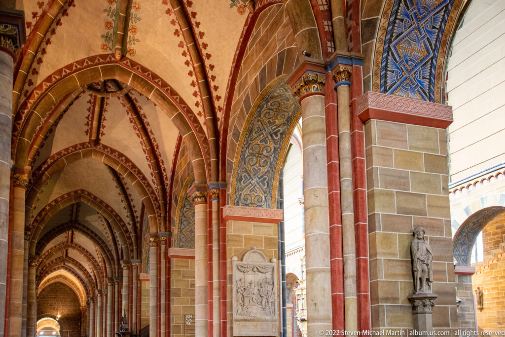 Long painted Gothic arcade with pointed vaults, red columns, decorative bands, memorial slabs, and statues.