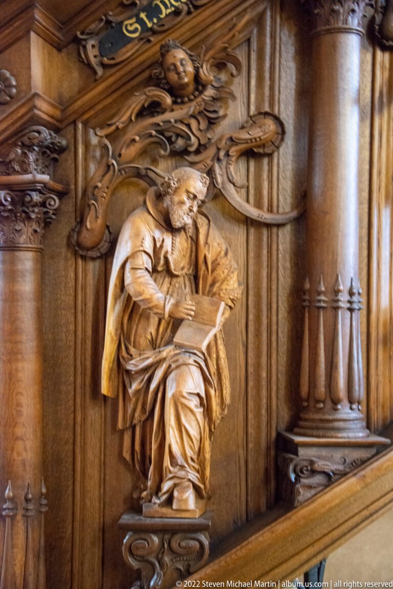 Carved wooden seated figure reading an open book beneath scrolling foliage and grotesque heads.