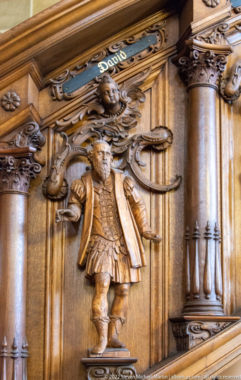 Standing carved male figure in period dress set within ornate wooden choir-stall architecture.