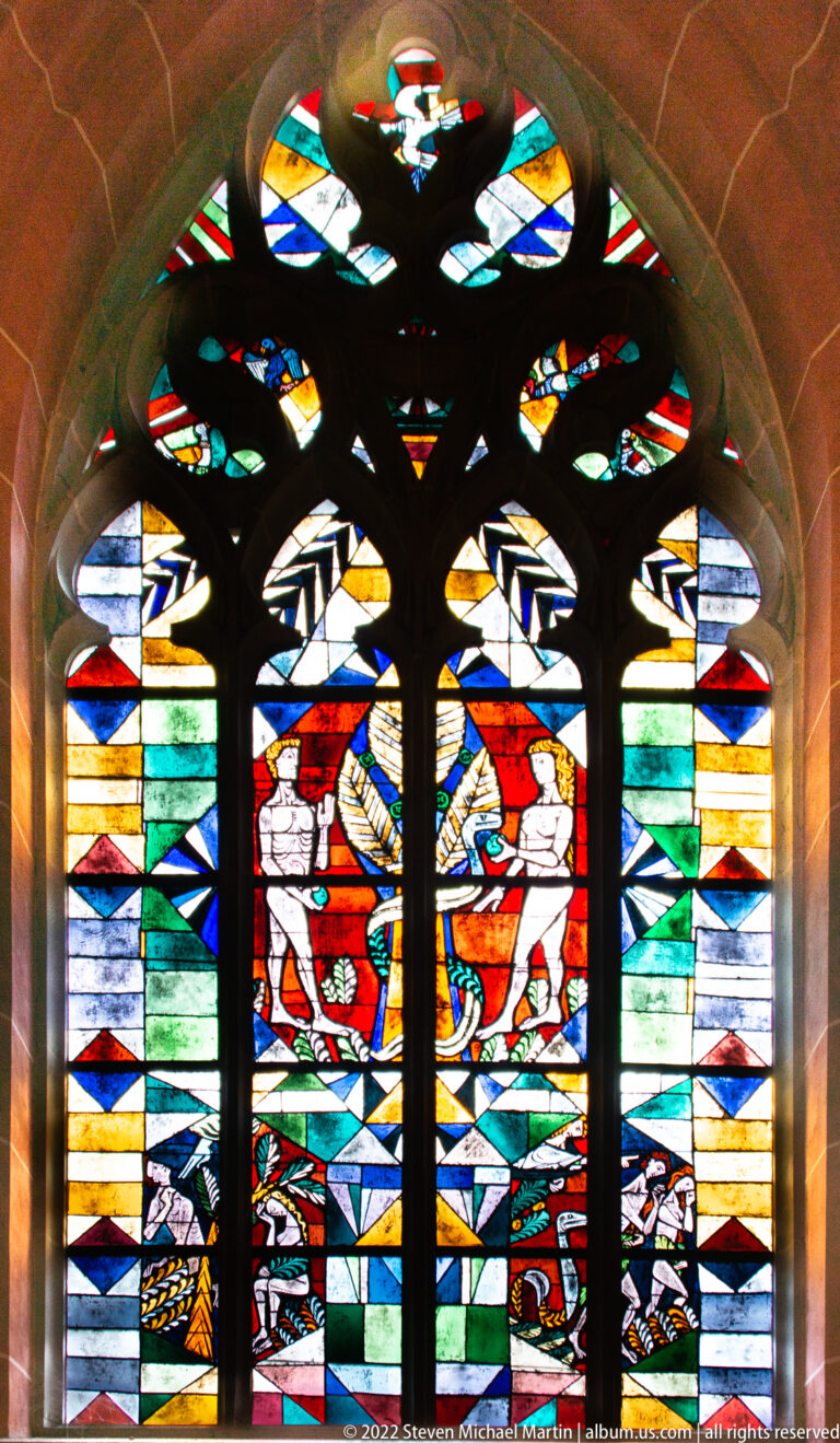 Broad stained-glass window with heraldic shields and central emblem beneath dark Gothic tracery.
