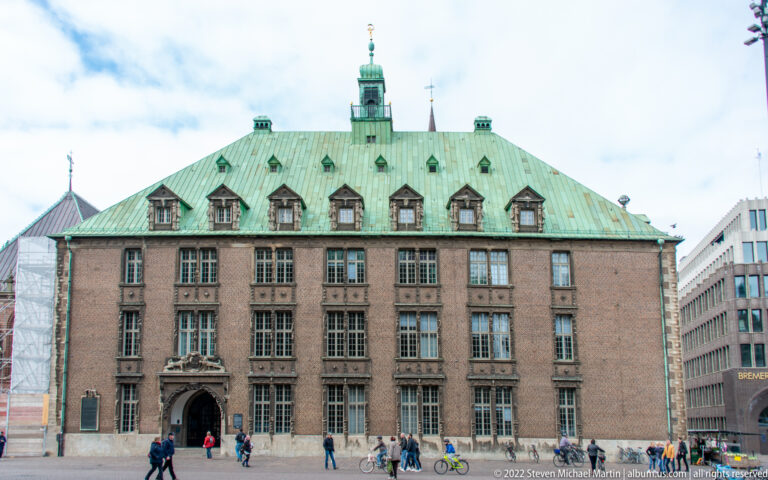 Large brick civic building with rows of windows, green copper roof, dormers, central entrance, pedestrians, and bicycles.