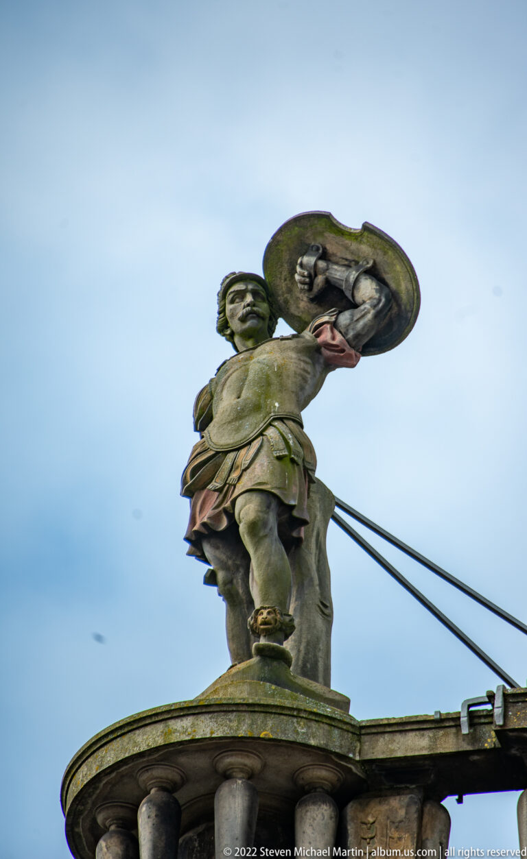 Painted roofline sculpture of a male figure holding a large brass instrument against the sky.