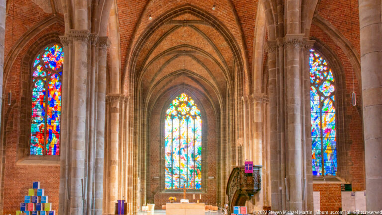 Brick-vaulted church interior with pointed arches, stone piers, altar, and colorful modern stained-glass windows.