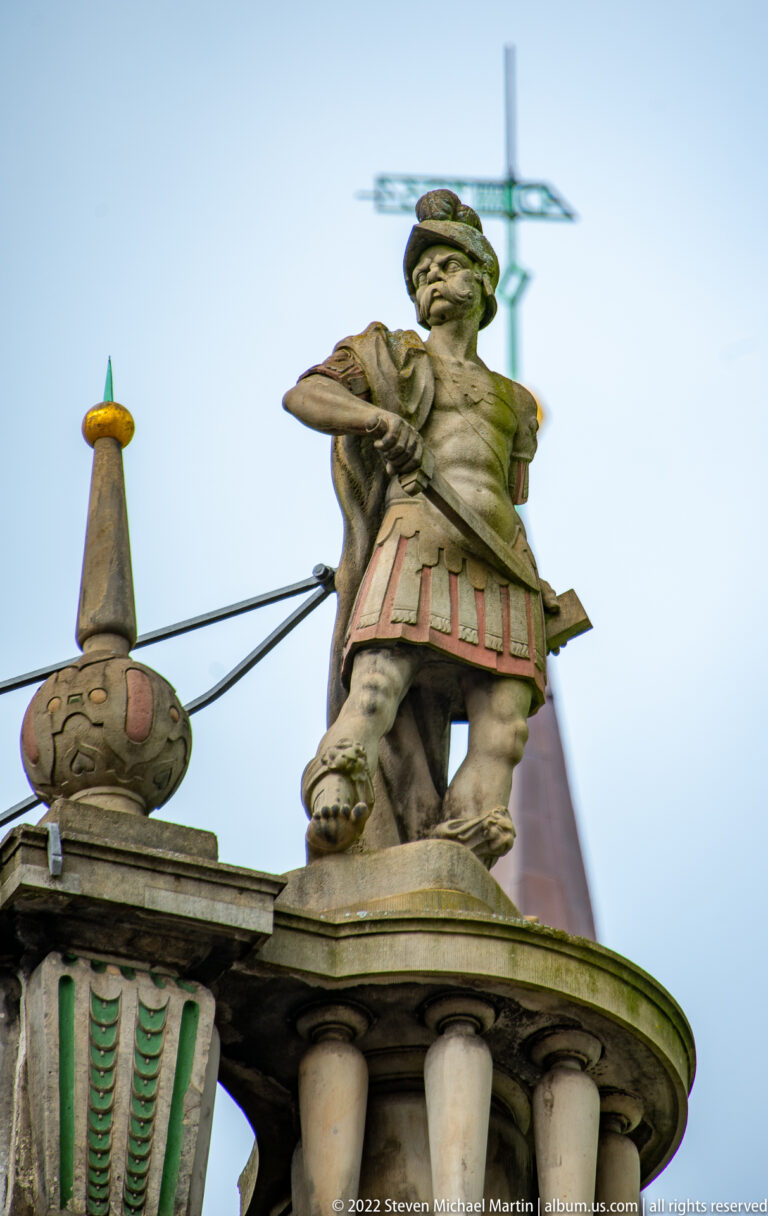 Painted rooftop sculpture of a male figure in historical costume standing on a balustrade.