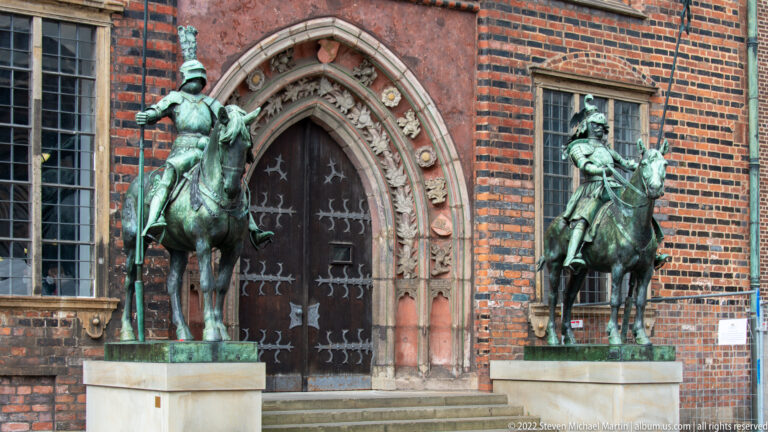 Two green-patinated equestrian bronze statues flank an ornate pointed-arch doorway in a red-brick historic building.