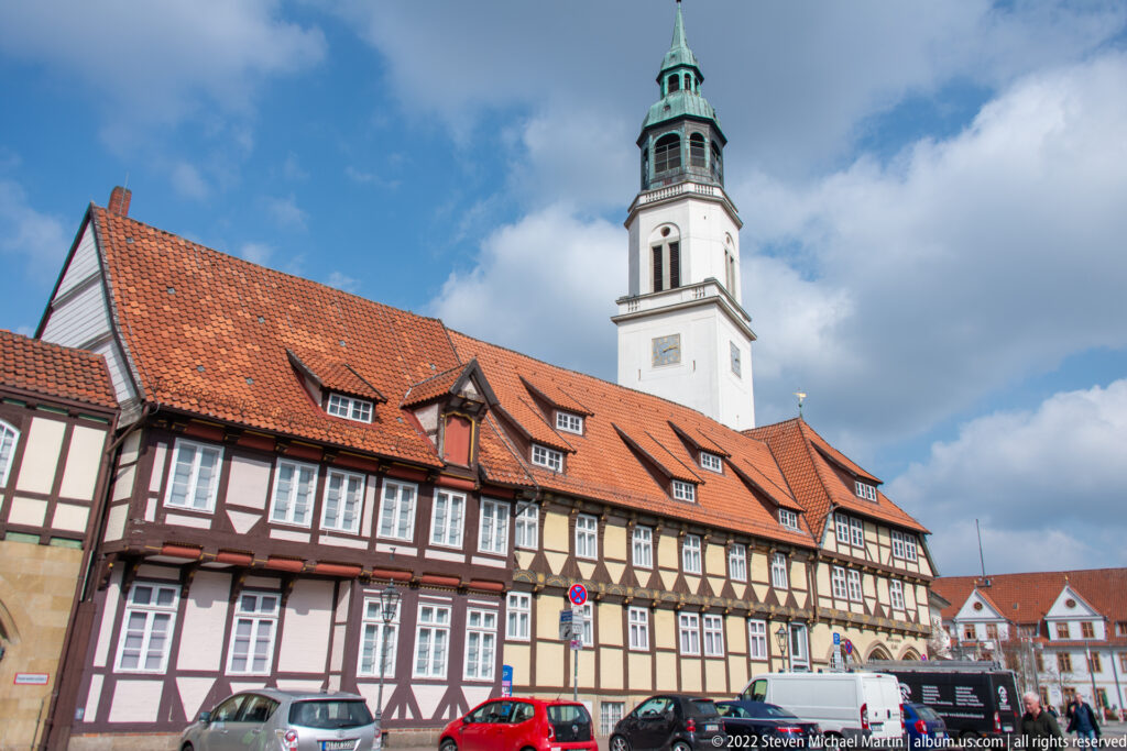 Long half-timbered building with red tile roof in front of a white church tower with green copper lantern.