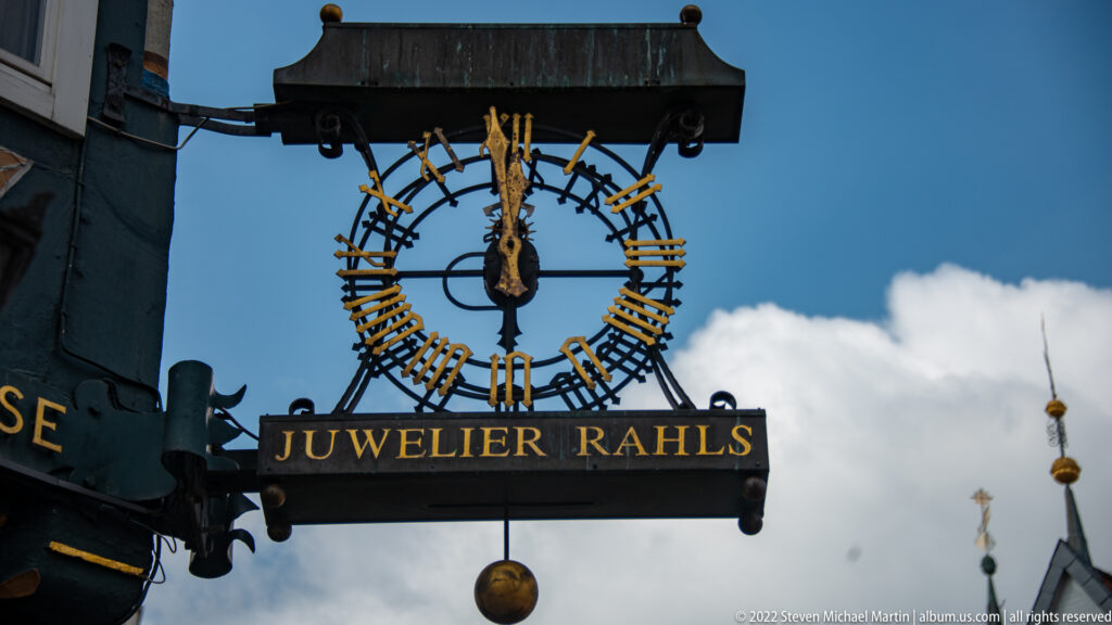 Ornate wrought-metal “JUWELIER RÄHLS” hanging sign with circular clock motif and suspended gilded sphere against blue sky.