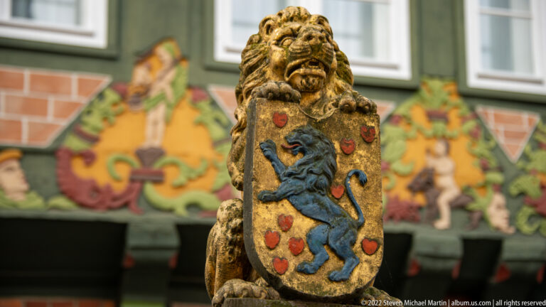 Carved lion holding a shield with a blue rampant lion and red motifs, set before colorful timber ornament.
