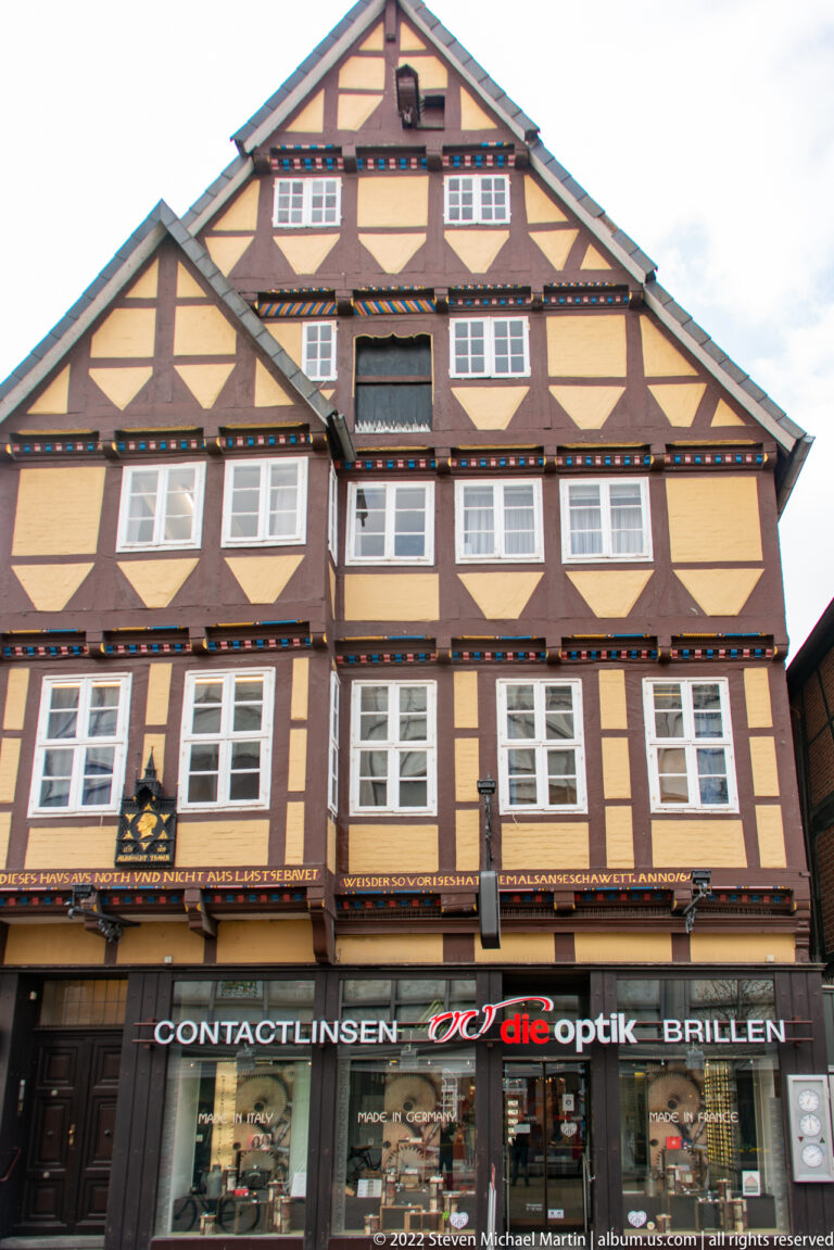 Broad half-timbered house with pale yellow panels and rows of white windows above a modern optical shop.