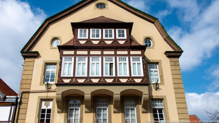 Pale gabled building with oval attic windows, a dark projecting timbered bay, and three arched openings below.