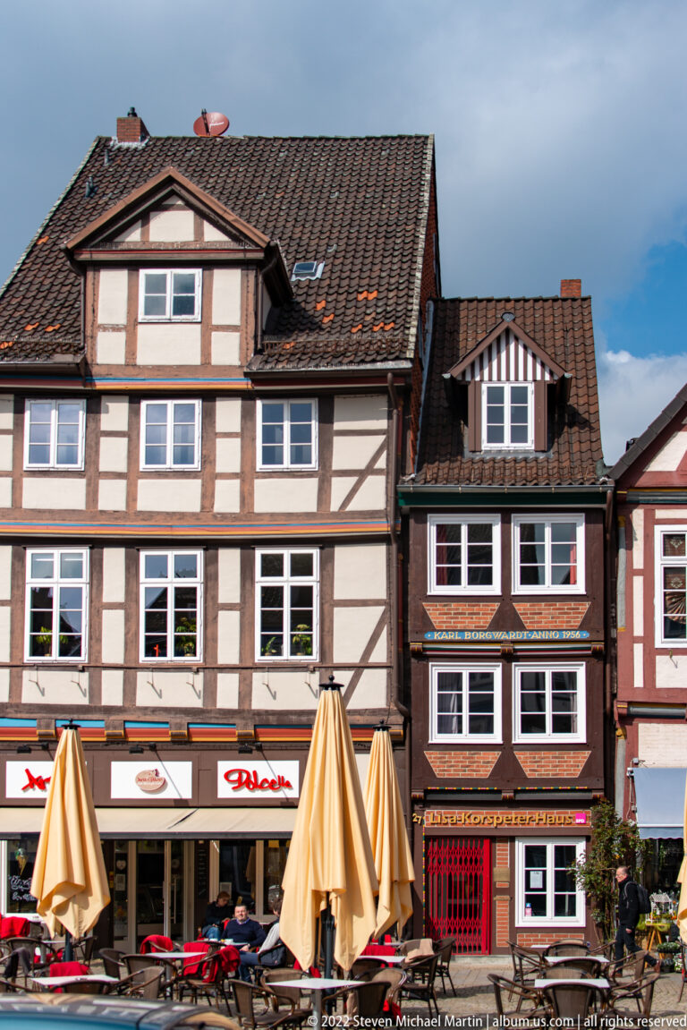 Two adjacent half-timbered houses with dormers above restaurant fronts, outdoor tables, and large umbrellas.