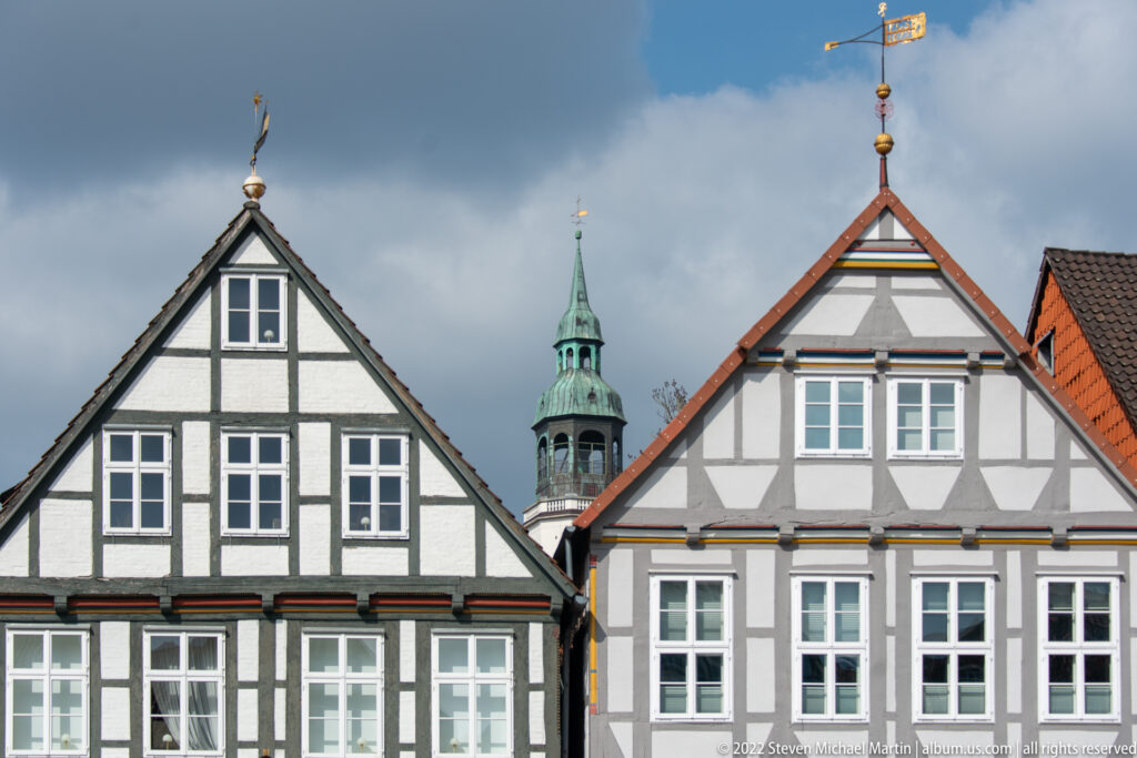 Two steep-gabled half-timbered houses with the green-topped church tower rising between them in the background.