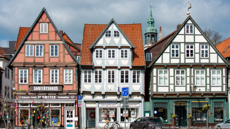 Pink, white, and green half-timbered houses with steep gables and modern shops along a Celle street.