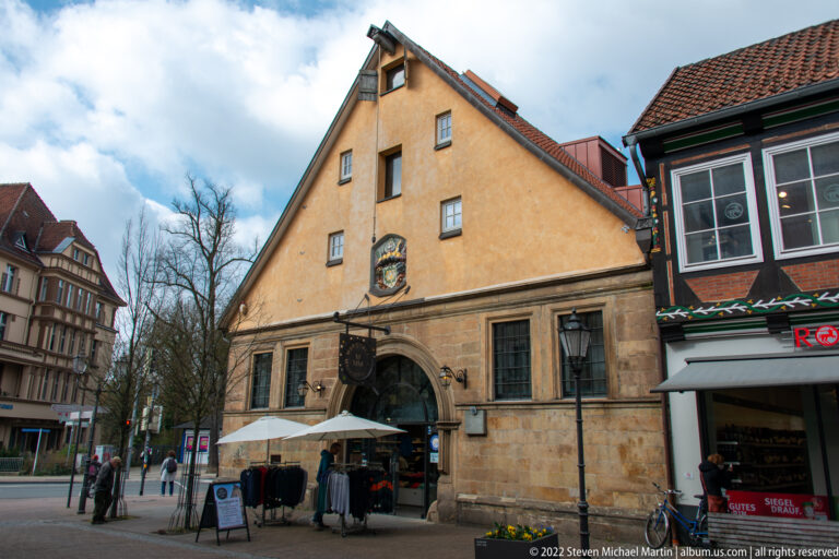 Ochre gabled building with stone lower façade, arched entrance, barred windows, and a heraldic plaque.