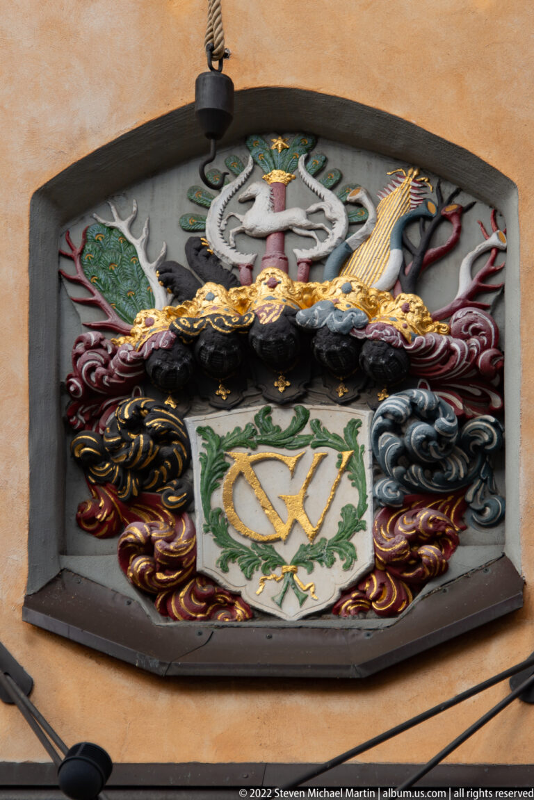 Colorful heraldic relief with white central shield, gold interlaced letters, scrollwork, and crest elements in an arched recess.