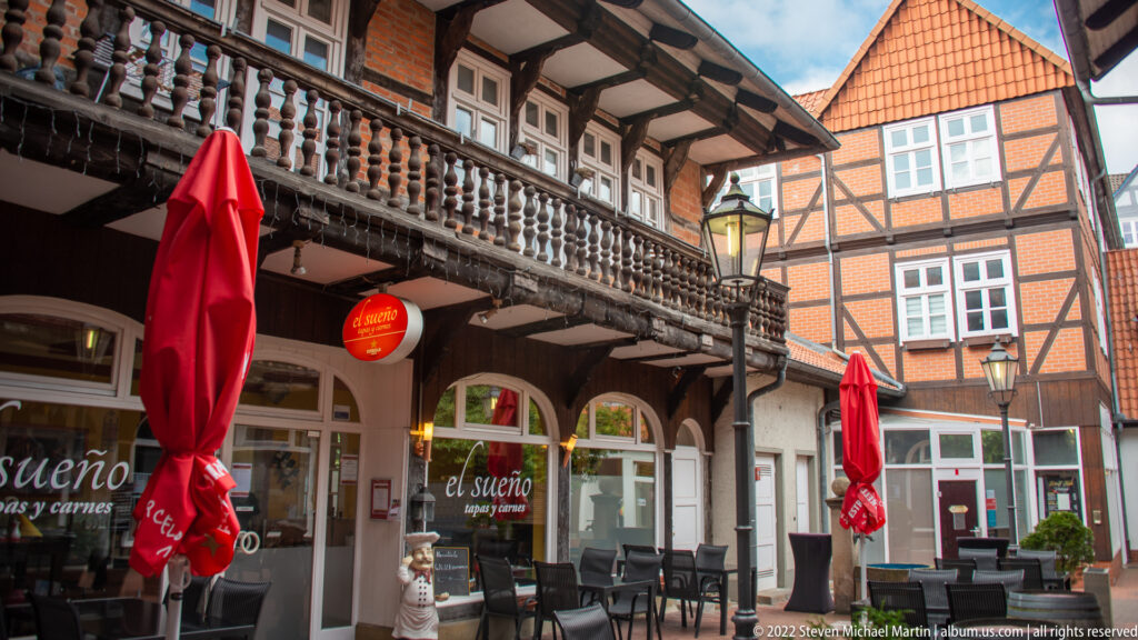 Historic courtyard with timber balcony, carved balusters, half-timbered walls, red café umbrellas, and outdoor tables.