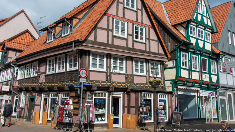 Pink half-timbered corner house with projecting upper floor beside a narrow green timber building and street-level shops.