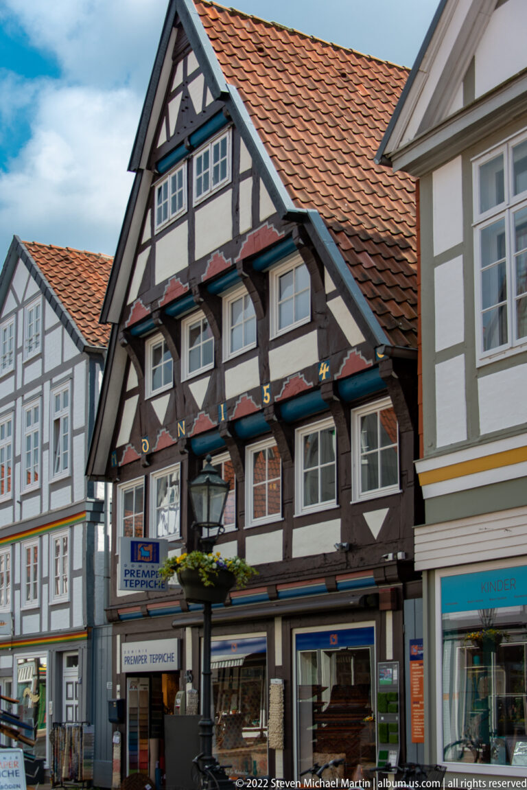 Dark half-timbered gabled house with small projecting upper windows, flower boxes, and modern shops at street level.