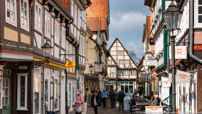 Narrow pedestrian lane in Celle lined with half-timbered shops, hanging signs, streetlamps, and people walking.