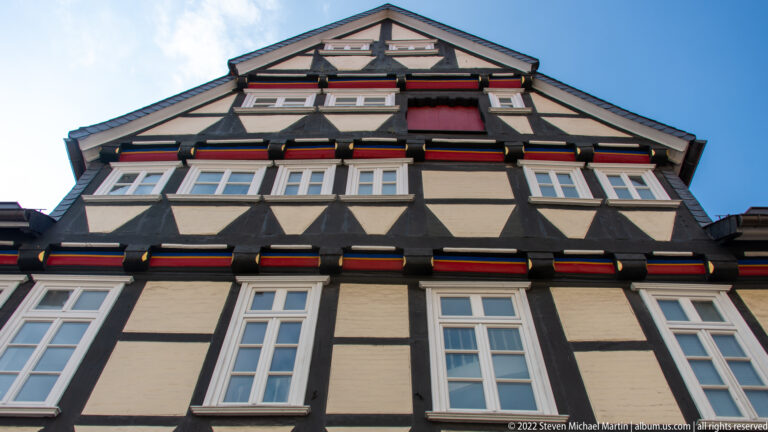 Upward view of a half-timbered gable with rows of white windows and red, yellow, and green painted beam ends.