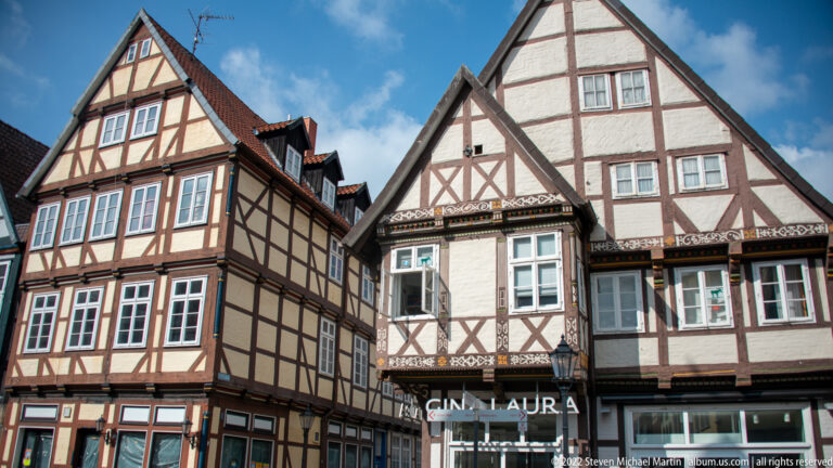 Two tall half-timbered gabled houses with decorative timber patterns and modern shopfronts, including a “GIN LAURA” sign.