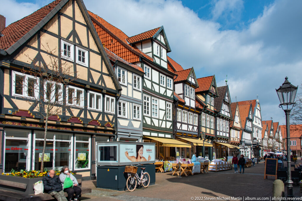 Long row of half-timbered houses along a pedestrian street with bicycles, outdoor displays, tables, and people.
