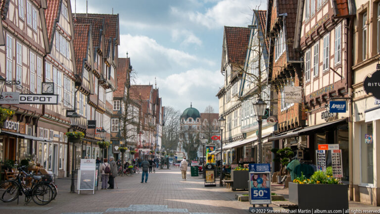 Pedestrian shopping street in Celle with historic façades, bicycles, signs, and a rounded roof visible in the distance.