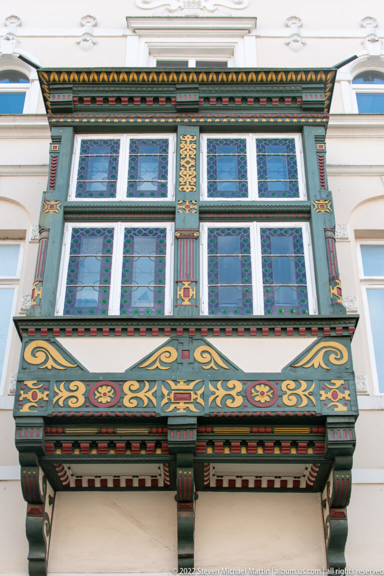 Close view of a green-painted projecting window bay with small-paned windows, colorful ornament, and bracketed supports.