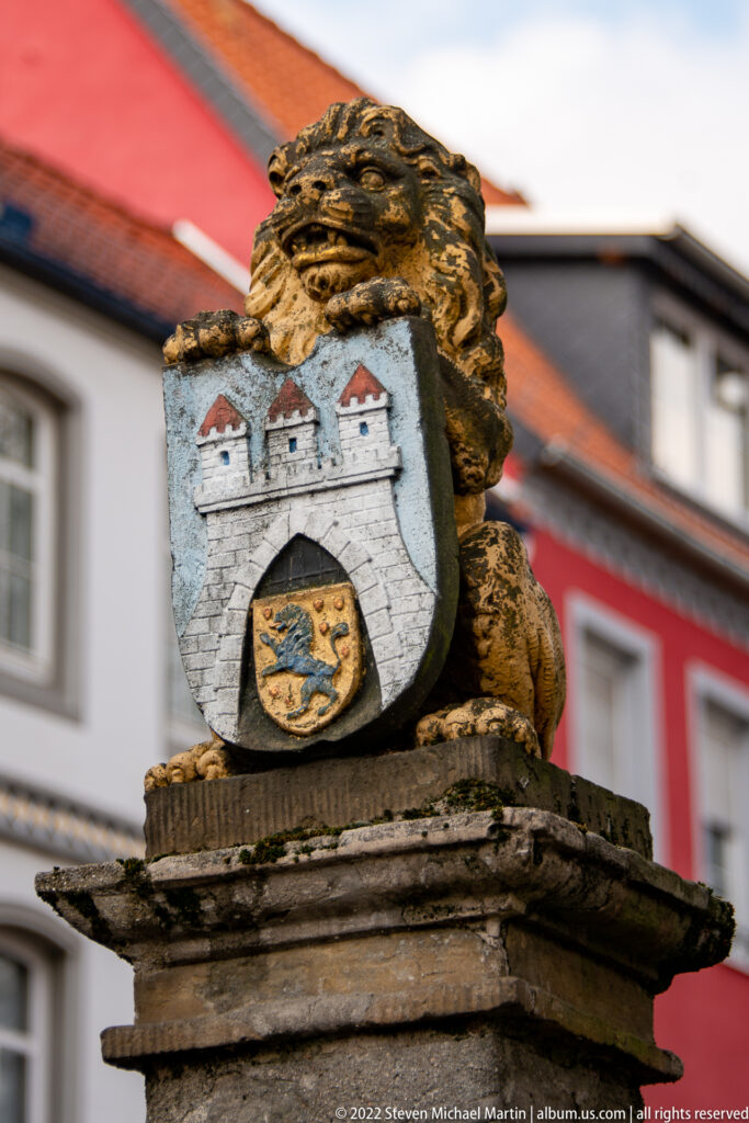 Weathered lion sculpture holding a painted heraldic shield, with red-roofed buildings softly blurred behind it.
